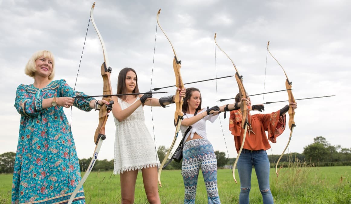 Four women practicing archery outdoors, holding bows and aiming in a grassy field under a cloudy sky.
