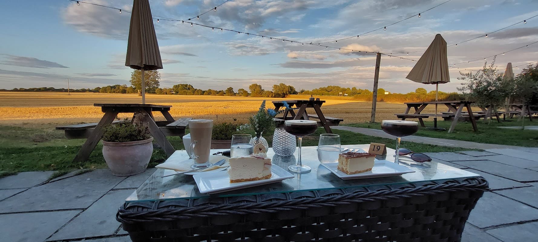 Desserts and drinks on a table with a scenic view of fields and trees under a sunset sky, patio umbrellas in the background.