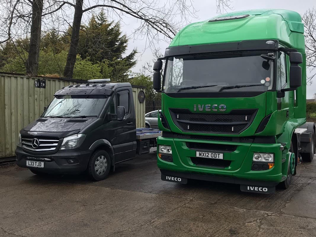 Two commercial vehicles parked side by side: a black Mercedes van and a green Iveco truck in a yard.