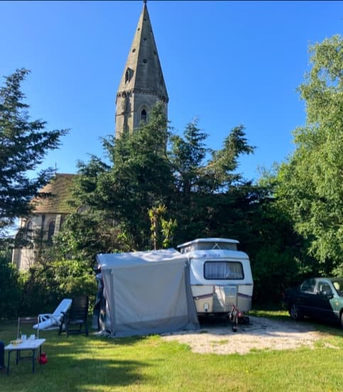 Caravan site with a camper, chairs, and a church tower in the background, surrounded by trees and blue sky.