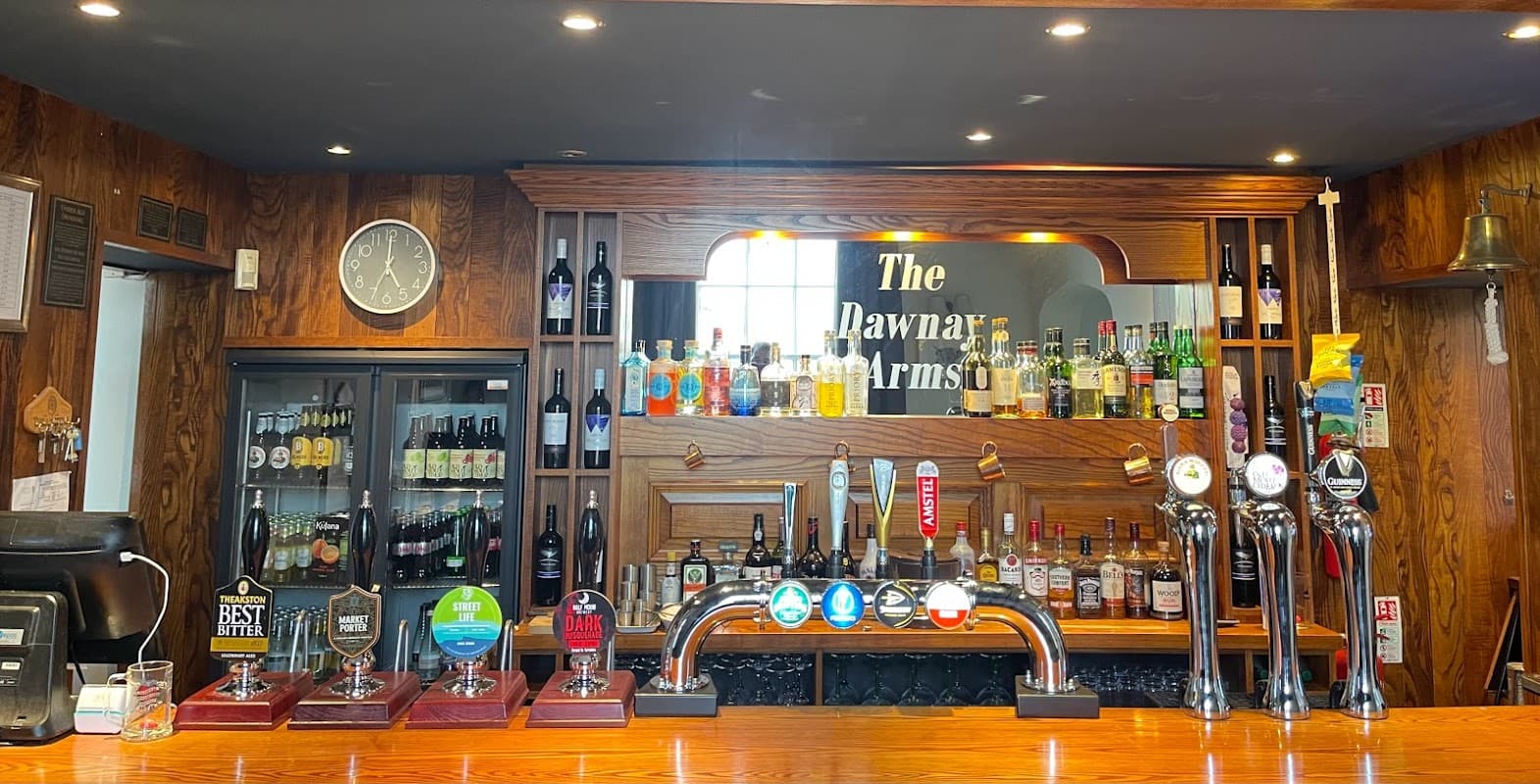 Wooden bar with a variety of beer taps, shelves of bottles, and a clock on the wall at The Dawnay Arms hotel.