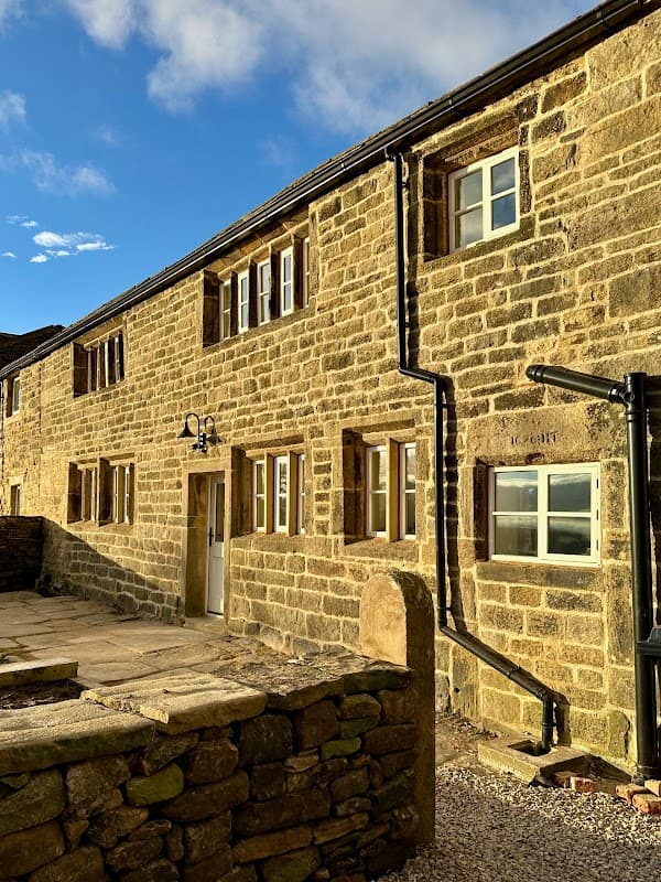 Stone cottage with multiple windows, set against a blue sky, featuring a paved area and stone wall in East Morton, Yorkshire.