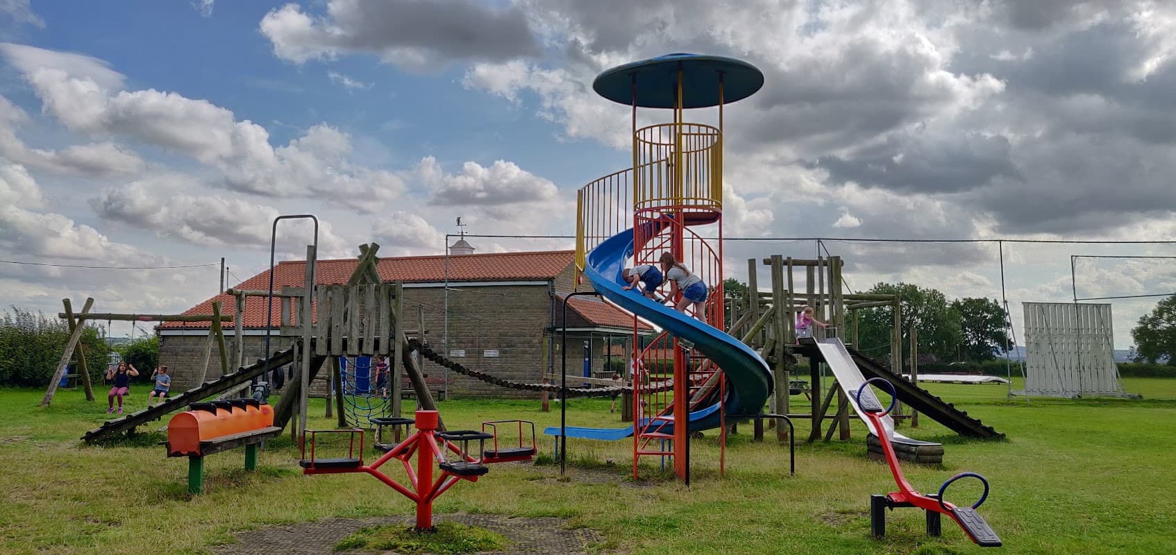 Colorful playground equipment, including slides and climbing structures, set on a grassy field under a cloudy sky.