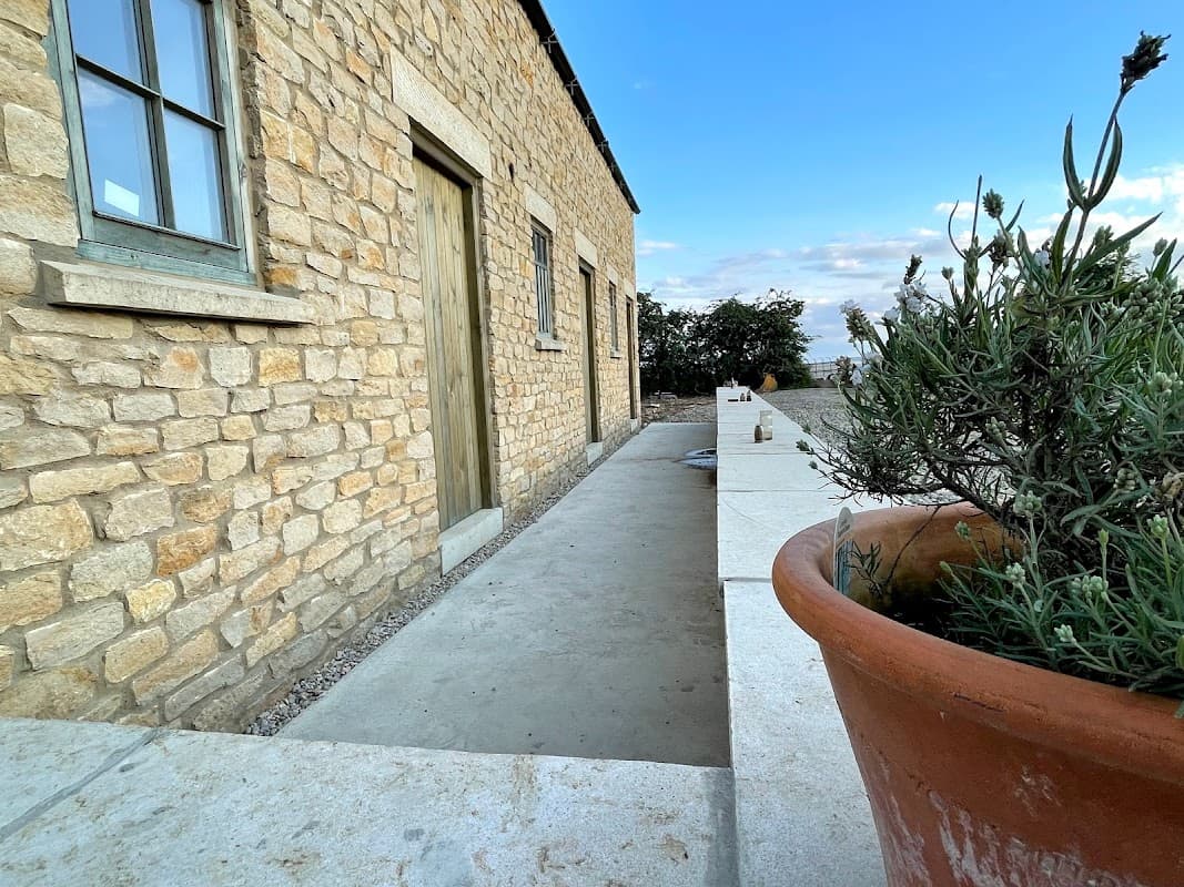 Stone building with a wooden door, a pathway, and a large terracotta pot with greenery under a blue sky.