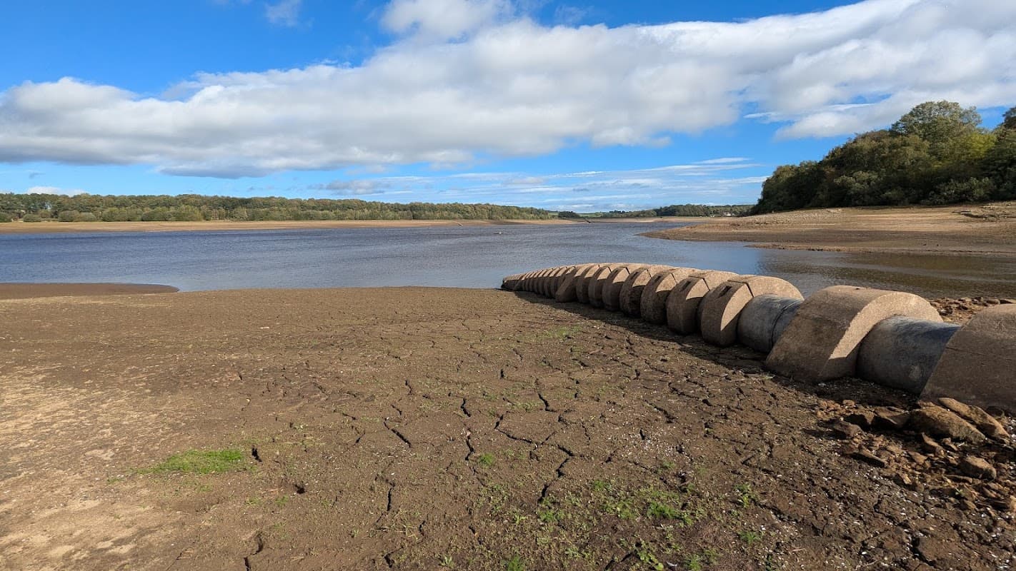 Dry shoreline of Eccup Reservoir with stone structures, blue sky, and scattered clouds in the background.