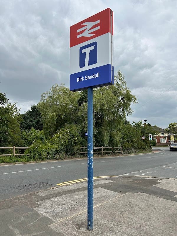 Kirk Sandall station sign on a pole near a car park, surrounded by greenery and cloudy skies.
