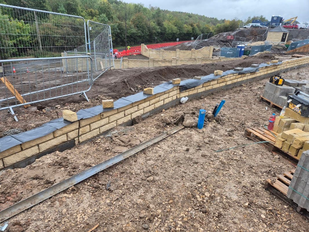 Construction site with a curved stone wall, gravel, and blue pipes, surrounded by fencing and building materials.