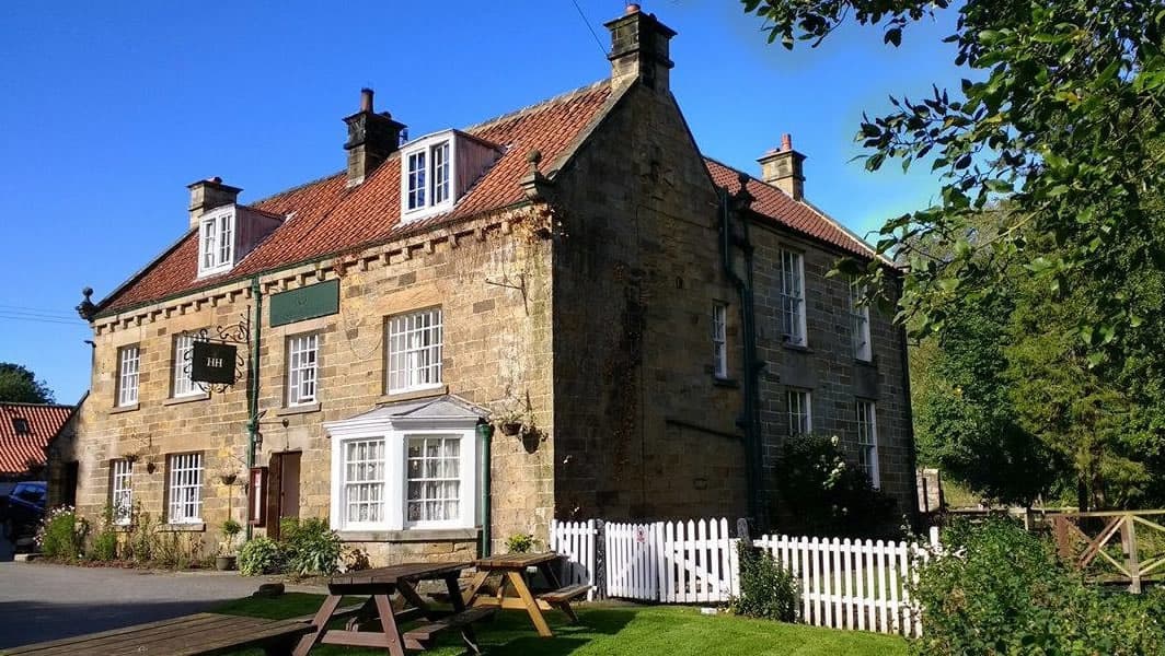 Historic stone pub with a red-tiled roof, white windows, and a green sign, surrounded by lush greenery and picnic tables.