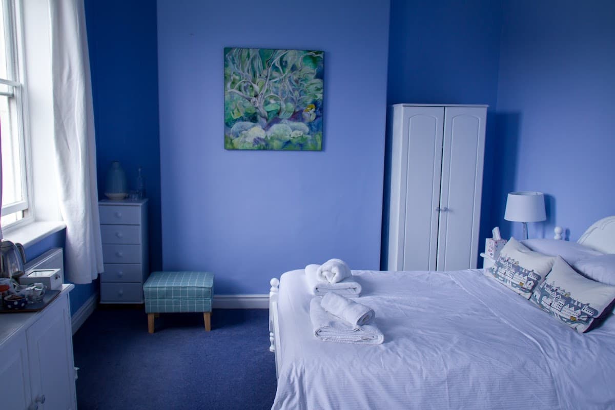 Cozy blue bedroom with a double bed, artwork on the wall, white furniture, and a small checkered stool.