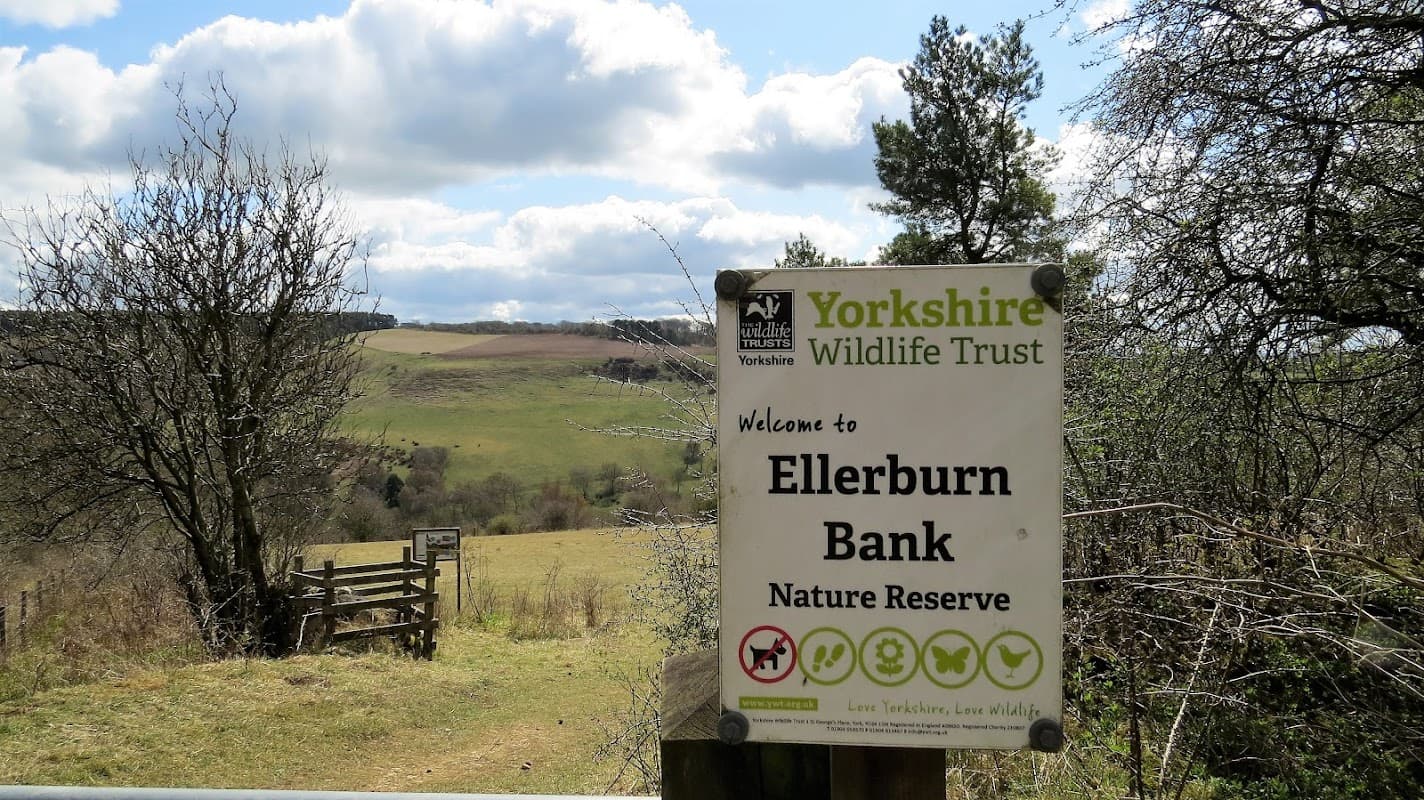 Sign for Ellerburn Bank Nature Reserve with rolling hills and trees in the background under a partly cloudy sky.