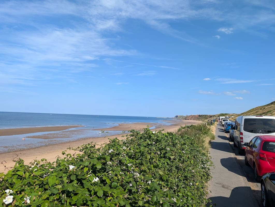 Car park lined with vehicles beside a sandy beach and calm sea under a clear blue sky in Whitby, Yorkshire.