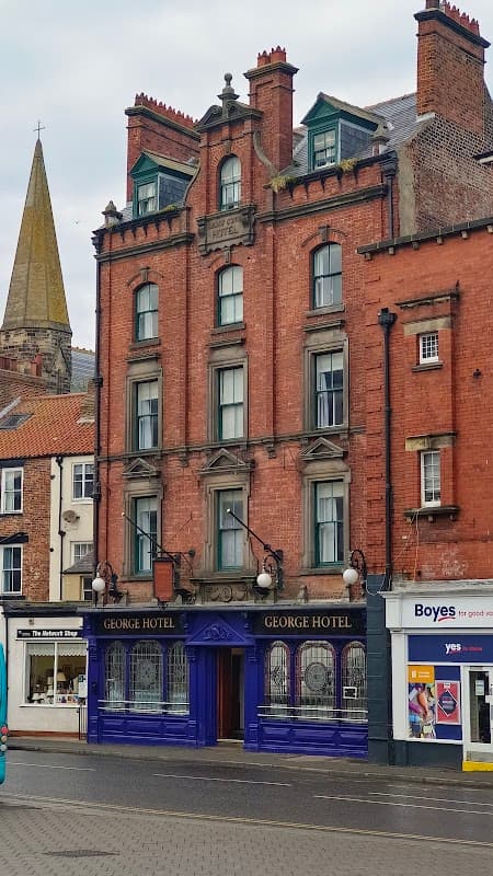 Victorian-style building with red brick facade, ornate details, and "George Hotel" sign; nearby shops and church spire visible.