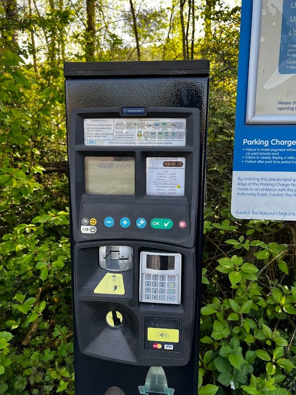 Parking meter with payment options, surrounded by greenery and a sign detailing parking charges and terms.