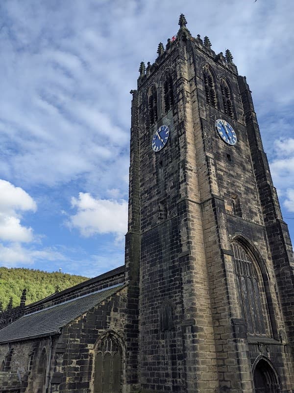 Historic stone clock tower with blue clock faces, surrounded by trees and a partly cloudy sky.
