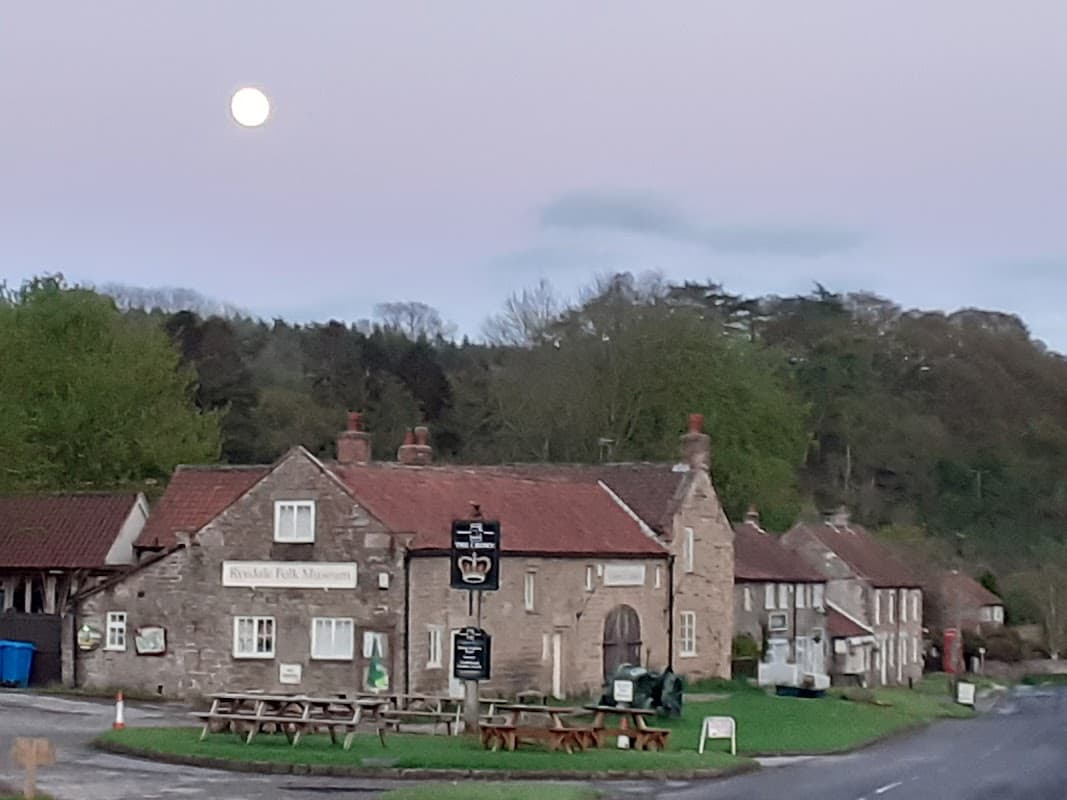 Ryedale Folk Museum building with stone walls, a sign, and outdoor seating, set against a twilight sky and trees.