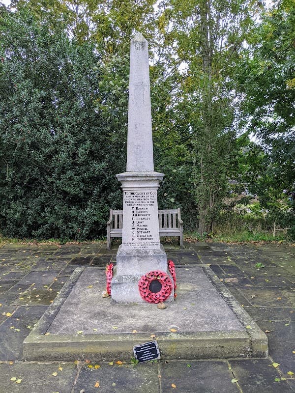 War memorial with an obelisk, surrounded by greenery and benches, adorned with poppy wreaths and a plaque.