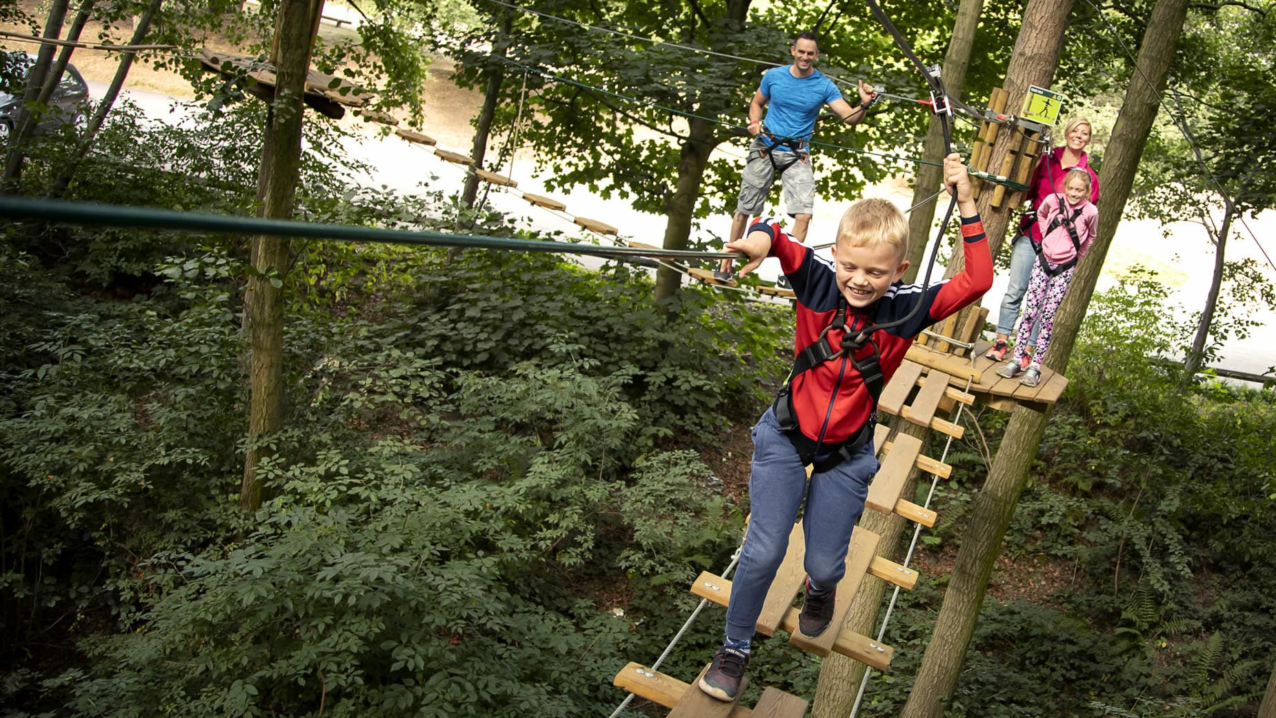 Child navigating treetop rope bridge in lush forest, with adults and other children in background, all wearing safety harnesses.