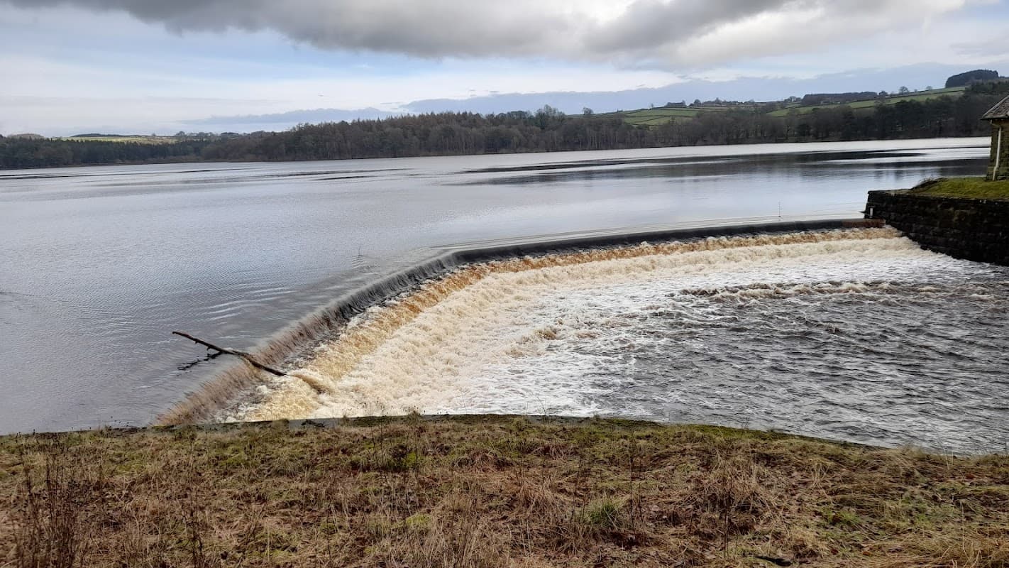 Water cascading over a stone dam into a calm reservoir, surrounded by trees and rolling hills under a cloudy sky.