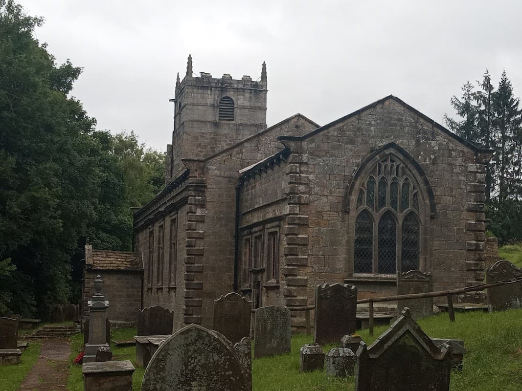 Historic stone church with gothic windows, surrounded by gravestones and lush greenery under a cloudy sky.
