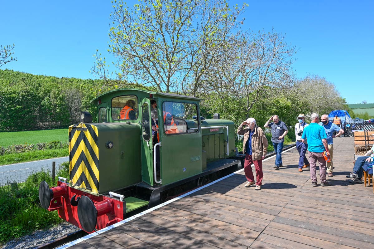 A green locomotive at Yorkshire Wolds Railway with visitors enjoying the sunny day and lush green fields in the background.