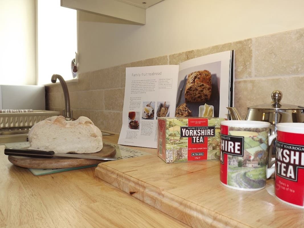 Freshly baked bread on a wooden board, with Yorkshire tea and a recipe book in a cozy kitchen setting.