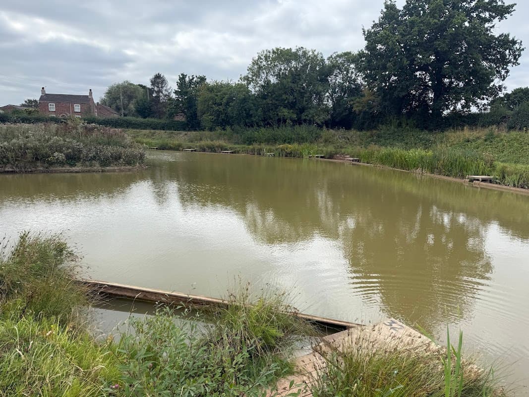 Calm pond surrounded by lush greenery and trees, with a distant house visible on the shore under a cloudy sky.