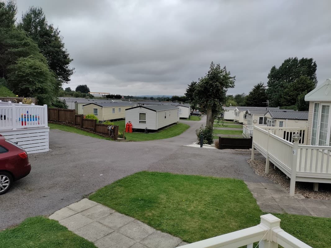 Caravans lined along a winding path, green lawns, and trees under a cloudy sky at Spring Willows Leisure Park.