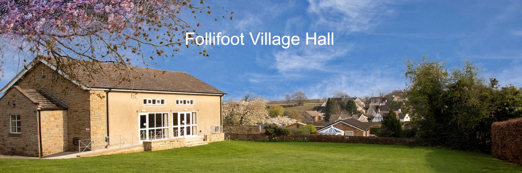 Follifoot Village Hall surrounded by greenery and houses, under a clear blue sky with blooming trees.