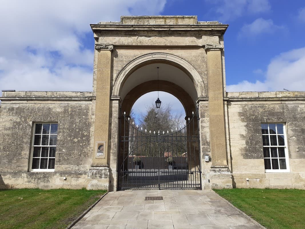 Historic stone archway with wrought iron gates, flanked by large windows and a grassy area under a blue sky.