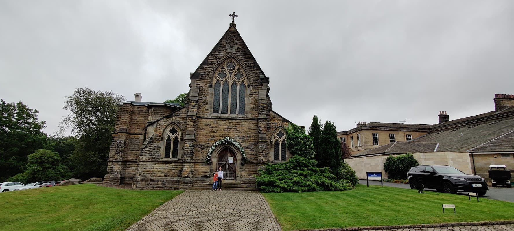 Victorian-style church with stone faΓ§ade, arched windows, and a cross atop, surrounded by greenery and a gravel path.