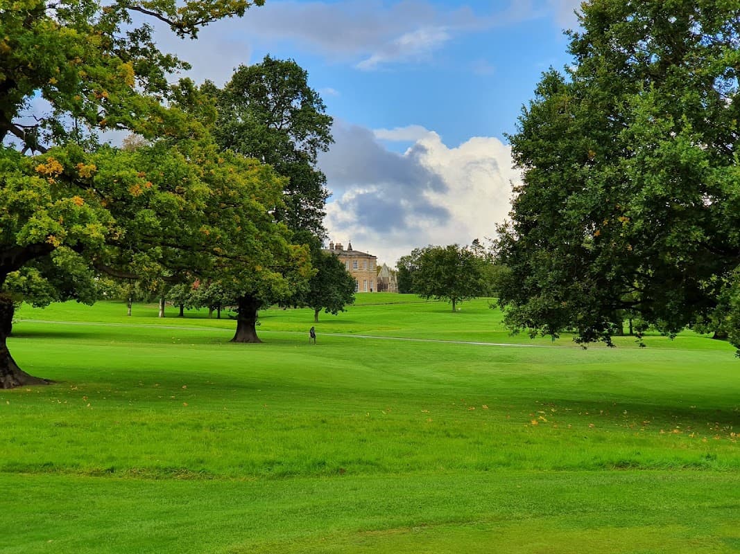 Lush green golf course with large trees and a historic building in the background under a partly cloudy sky.