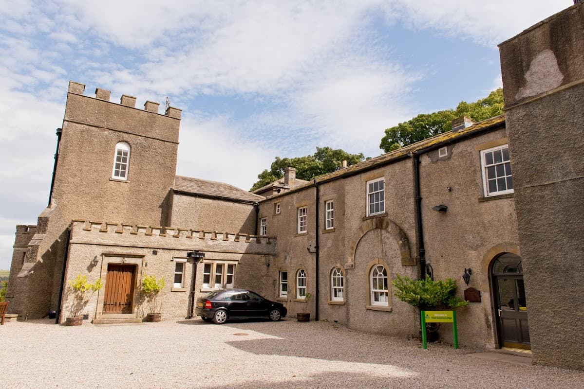 Historic stone building with a tower, arched windows, and a gravel parking area surrounded by greenery.