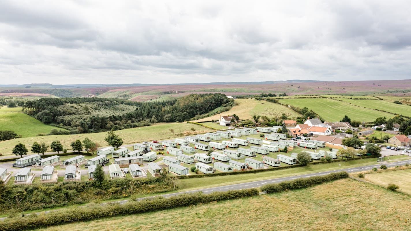 Aerial view of The Flask Holiday Park with green caravans, fields, and hills in the background under a cloudy sky.