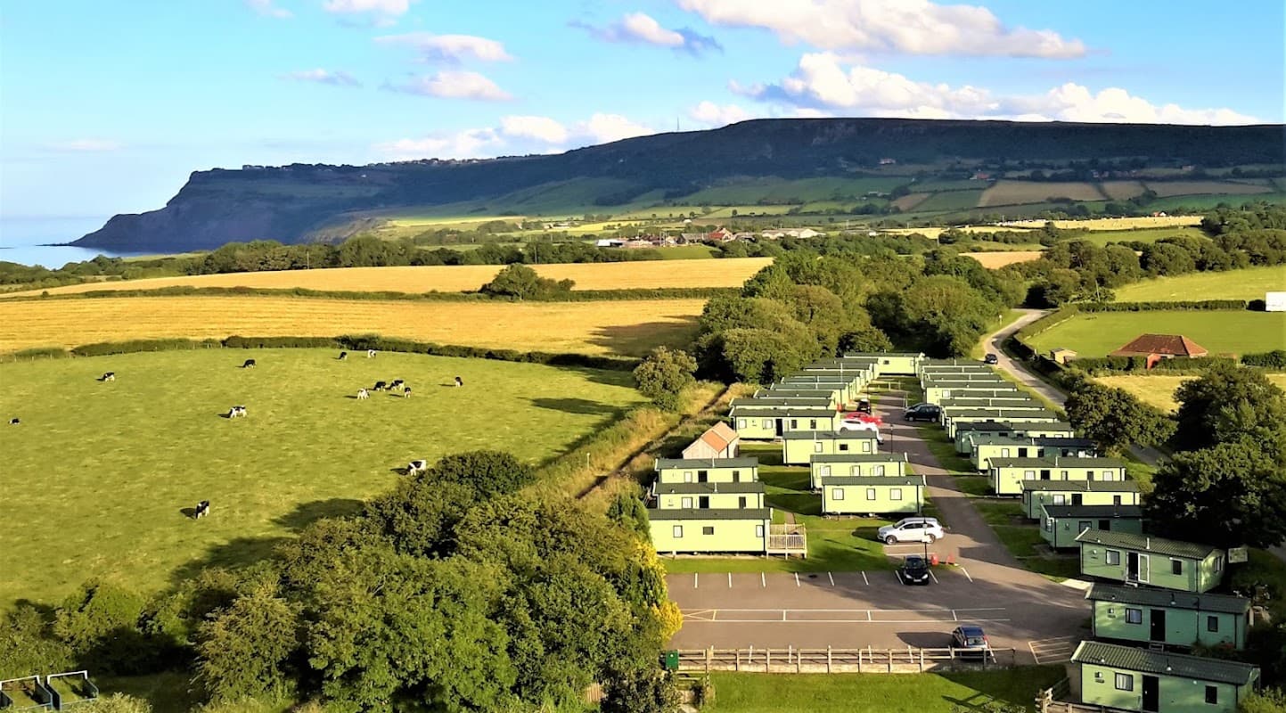 Campsite with green lodges, grassy fields, and distant hills overlooking the sea in Fylingthorpe, Yorkshire.