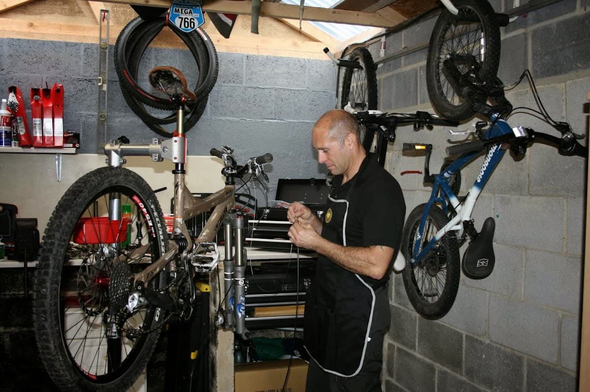 A mechanic repairs a bike in a workshop, surrounded by tools, bike parts, and two mounted bicycles on the wall.