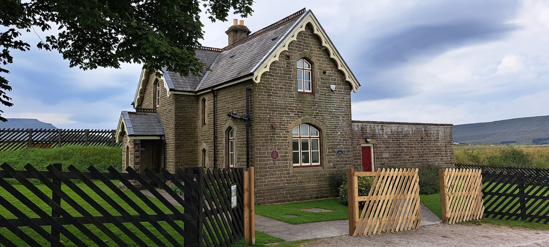 Historic stone building with a gabled roof, surrounded by a wooden fence and green grass, set against a cloudy sky.