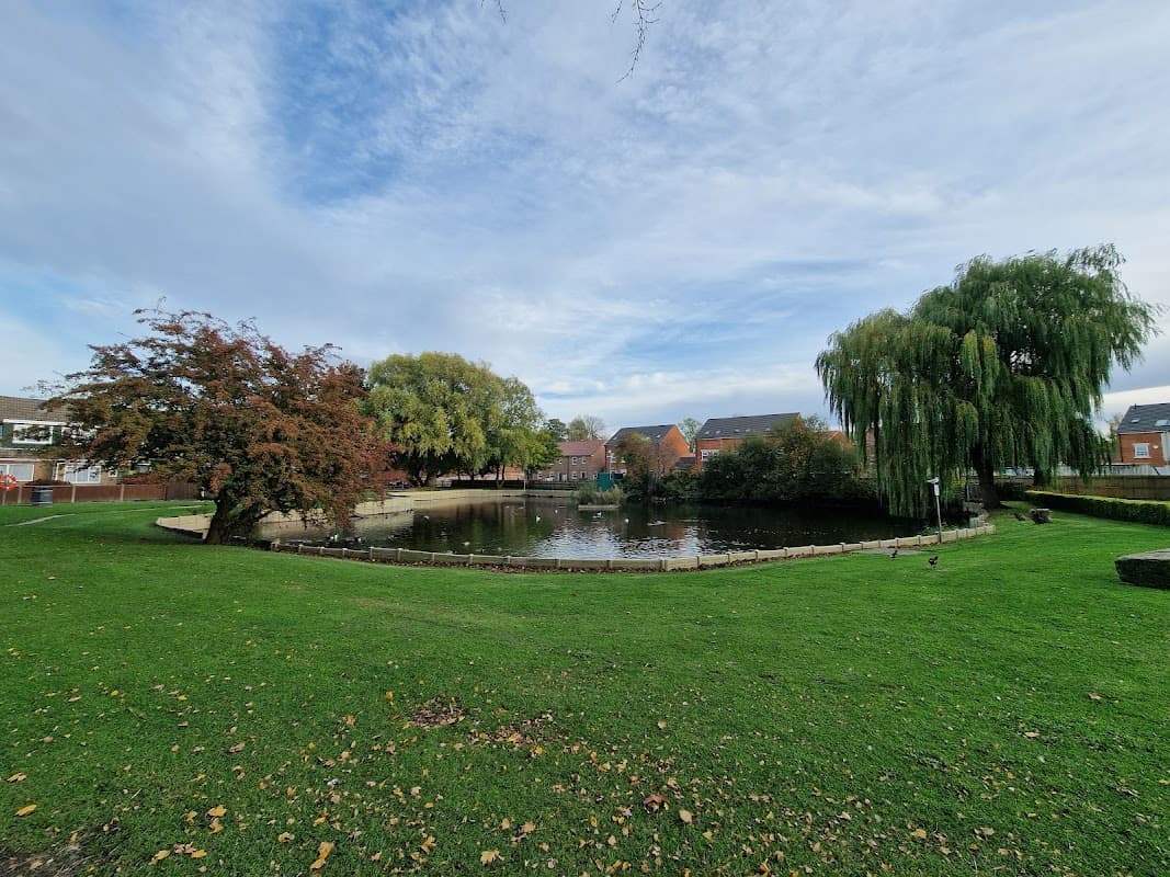 Lush green park featuring a serene pond surrounded by trees and residential buildings under a cloudy sky.