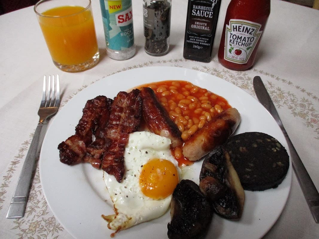 A hearty breakfast plate with bacon, sausages, baked beans, a fried egg, black pudding, and mushrooms, alongside sauces.