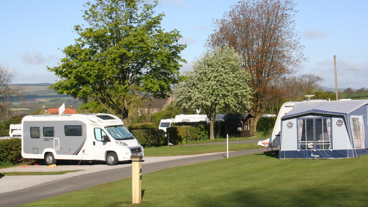 Caravans and motorhomes parked on a grassy campsite with trees and a clear blue sky in Gilling West, Yorkshire.