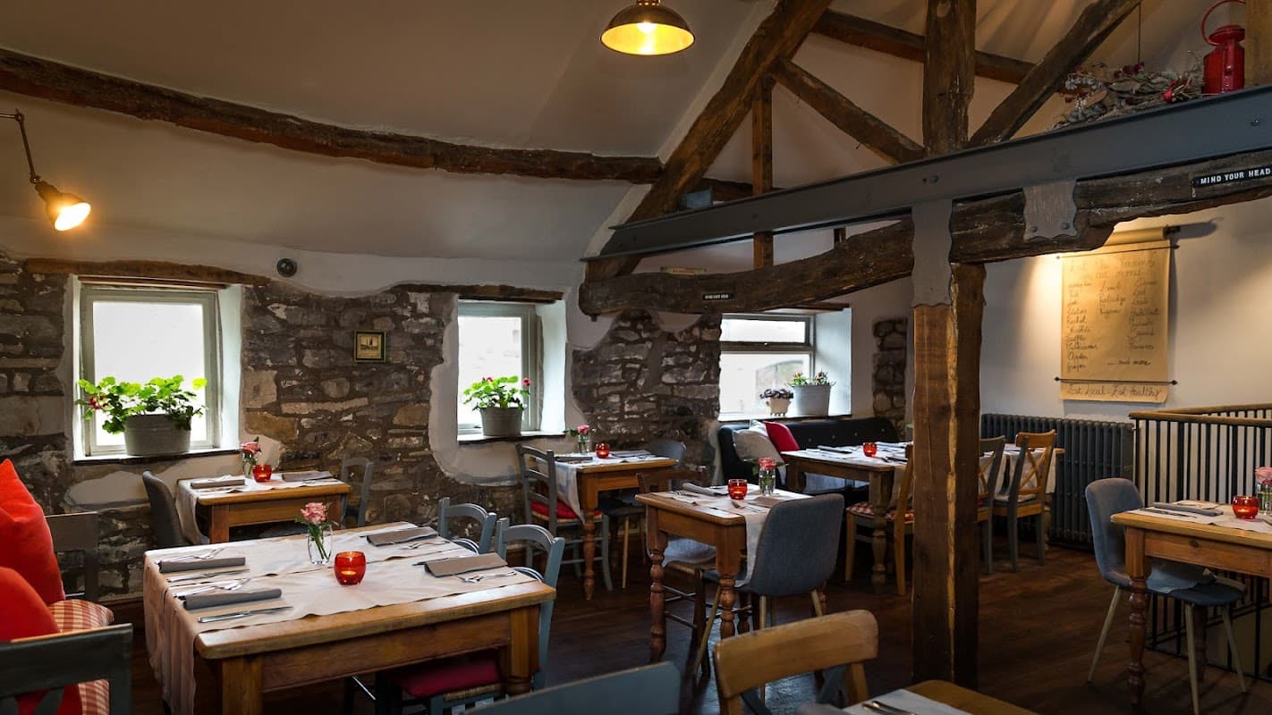 Rustic dining area with wooden beams, stone walls, tables set with cutlery, flowers, and warm lighting.