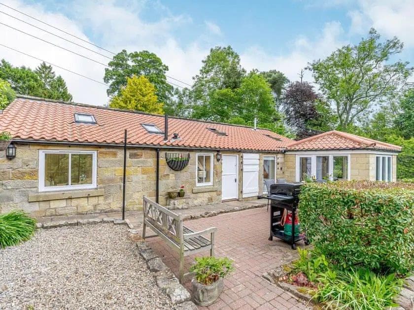 Charming stone cottage with a red-tiled roof, surrounded by greenery and a paved patio area with outdoor seating.