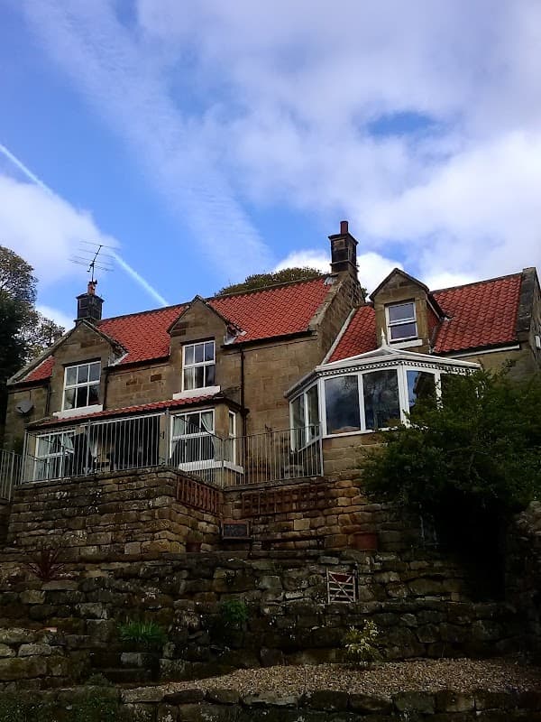 Stone house with red-tiled roof, large windows, and a garden, set against a blue sky with wispy clouds.