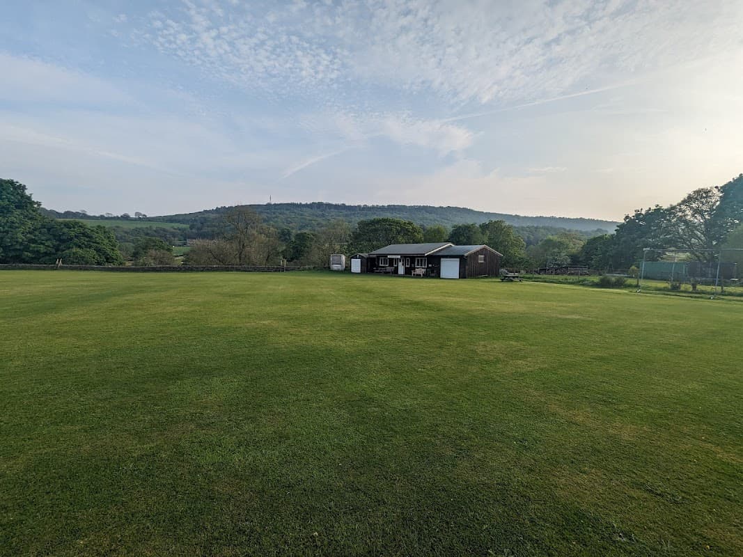 Green cricket field with a clubhouse and trees, set against a backdrop of hills and a clear blue sky.