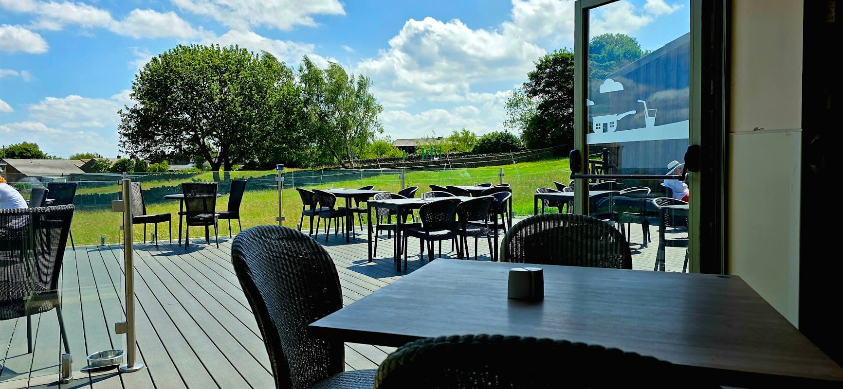 Outdoor seating area with black chairs and tables, overlooking green fields under a blue sky with fluffy clouds.