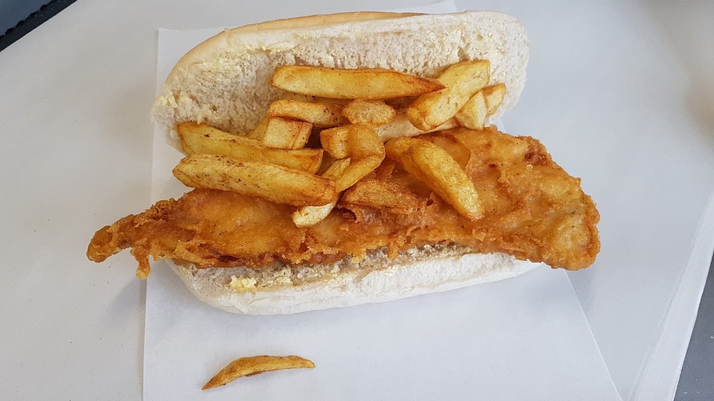 Crispy battered fish fillet and golden chips served in a soft bread bun on a white paper surface.