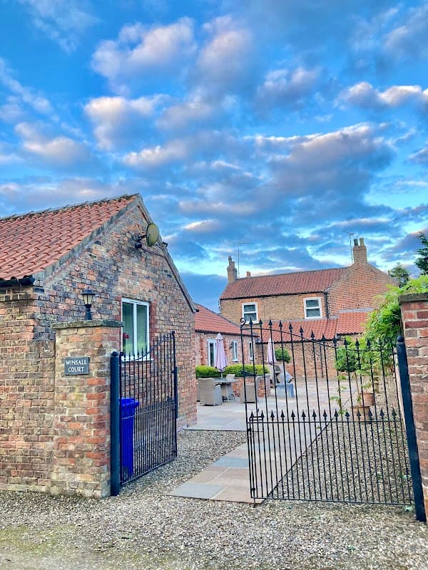 Rustic brick buildings with a gated entrance, under a blue sky with clouds, in a tranquil countryside setting.