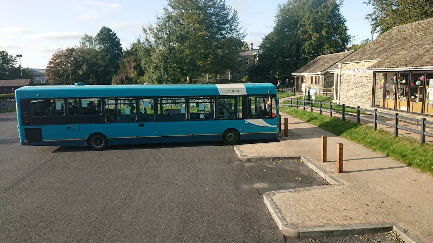 A teal bus parked near a stone building with large windows, surrounded by trees in a rural setting.