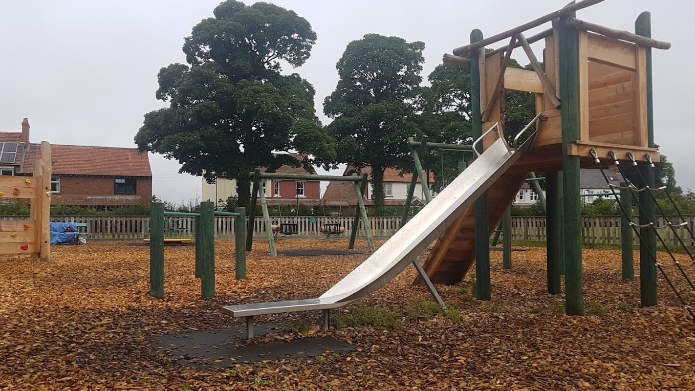 Playground with a wooden slide, climbing frame, and surrounding trees, covered in autumn leaves.