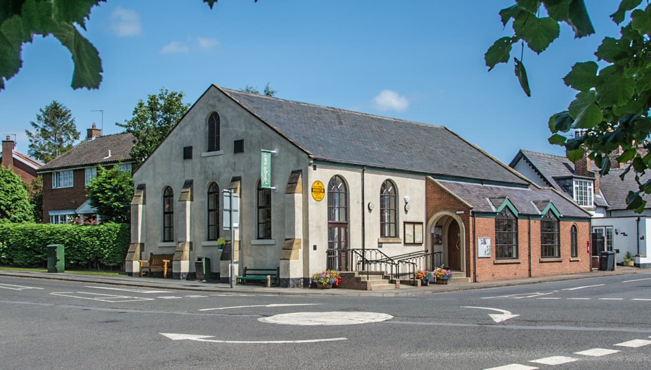 Great Broughton Village Hall with a stone facade, large windows, and surrounding greenery on a sunny day.