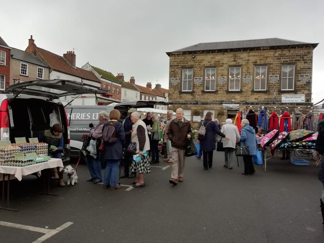 Busy market scene with people browsing stalls, a van, and colorful clothing hanging nearby in Great Broughton.